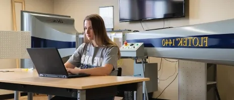 A female engineer sitting at a computer with a large wind tunnel in the background.